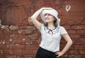 A smiling young woman in a white blouse and a hat posing against the backdrop of an old vintage brown brick wall. Female hand holds a hat. Photos in retro style.
