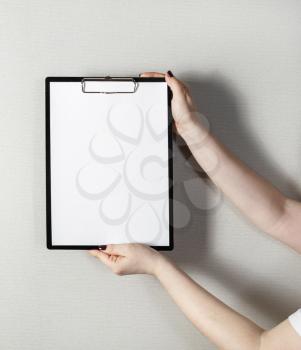 Clipboard with a blank sheet of paper in female hands with a deep shadow on the background of gray wall. Mock-up for design presentations and portfolios.