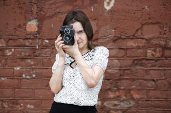 Girl with an old vintage camera on a brick wall background. Young woman photographer taking photo outdoor. Selective focus on model. Retro style photo.