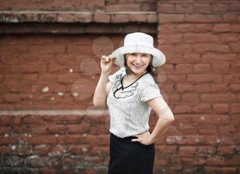 A smiling young woman in a white blouse and a hat posing against the backdrop of an old vintage brown brick wall. Female hand holds a hat.  Photo in retro style. Selective focus on model.