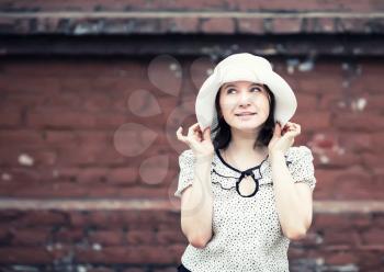Pretty young smiling woman in a white blouse and hat posing on old vintage brick wall background. Girl holds the edges of the hat by hand. Toned photo with copy space. Vintage style photo.
