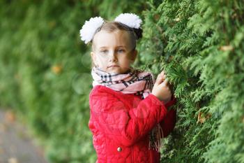 Portrait of a serious little baby girl in a red jacket and scarf on a blurred background of green bushes. Shallow depth of field. Selective focus.