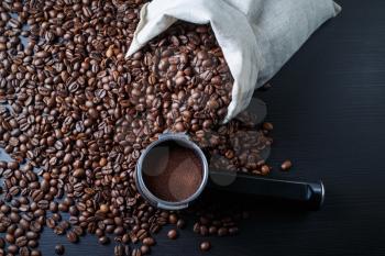 Coffee beans scattered from a canvas bag and holder from an espresso machine against a black wooden table background.