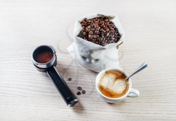 Coffee cup, holder from an espresso machine and coffee beans in a canvas bag on light wooden oak table background. Still life with coffee.