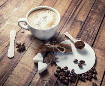 Coffee cup, cinnamon sticks, coffee beans, anise, sugar, spoon and coasters on vintage wooden kitchen table background.