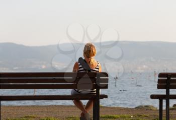 Lonely young woman sitting on a wooden bench and looking at the sea.