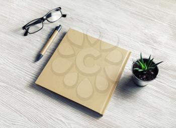 Closed blank notepad, glasses, pen and plant on light wooden background. Blank stationery set.