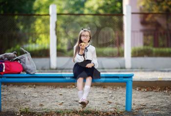 Schoolgirl in school uniform eats an apple. Child girl sitting on a bench. Selective focus.