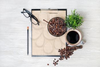 Stationery set and coffee. Clipboard with kraft sheet of paper, coffee cup, coffee beans, pencil, glasses and plant on light wooden background. Flat lay.