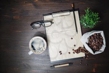 Coffee and kraft stationery. Clipboard with blank vintage letterhead, coffee cup, coffee beans, glasses, pencil and plant on wood table background. Flat lay.