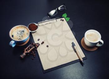Blank retro stationery set and coffee on black table background. Clipboard with blank kraft letterhead, coffee cups, coffee beans, pen, glasses and ground powder.