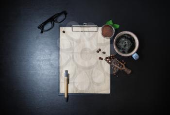 Blank vintage stationery set and coffee on black table background. Clipboard with blank kraft letterhead, coffee cup, coffee beans, pen, glasses and ground powder. Top view. Flat lay.