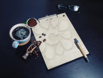 Clipboard with blank kraft paper, coffee cup, coffee beans, glasses and pen on black wood table background.