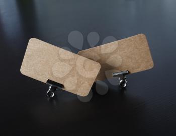 Two blank kraft business cards and metal binder clips on black wooden background. Mockup for branding identity.