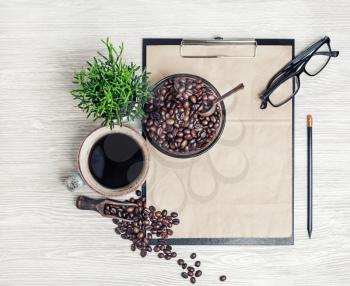Clipboard with blank kraft letterhead, coffee cup, coffee beans, pencil, glasses and plant on light wood kitchen table background. Top view. Flat lay.
