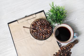 Still life with coffee cup, clipboard with kraft paper, plant and coffee beans on light wooden background.