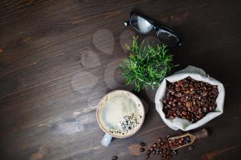Delicious fresh coffee. Coffee cup, coffee beans in canvas sack, plant and glasses on wood table background. Top view. Flat lay.