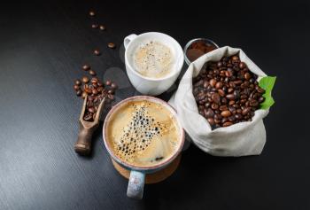 Still life with coffee cups and coffee beans in canvas bag on black kitchen table.