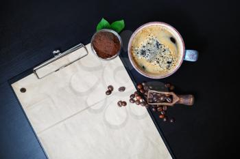 Photo of coffee cup and stationery. Clipboard with blank kraft letterhead, coffee beans and ground powder on black table background. Top view. Flat lay.