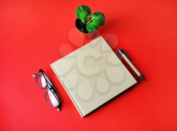 Blank square kraft book, glasses, pen and plant on red paper background. Template for placing your design.
