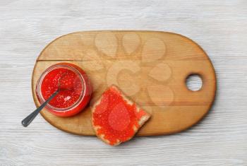 Sandwich with red caviar, glass jar and spoon on wooden cutting board. Top view. Flat lay.
