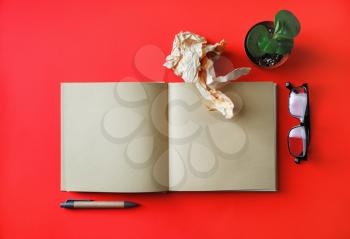 Blank kraft booklet, plant, pen, glasses and crumpled paper on red paper background. Responsive design mockup. Flat lay.