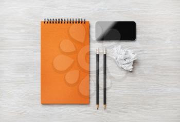 Blank orange notepad, smartphone, pencils and crumpled paper on light wood table background. Top view. Flat lay.