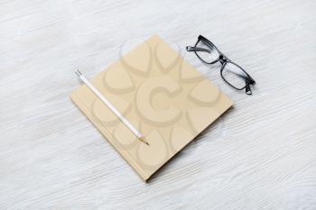 Photo of closed blank square book, glasses and pencil on light wooden background. Template for placing your design.