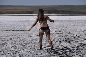 Woman walking along the dry bottom of a salt lake, rear view. Walk the dark-haired woman in a swimsuit on the bottom of a dry lake with salt and mud. The ancient dried-up lake.