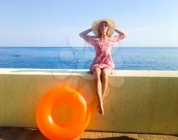 A woman with a beautiful figure is sitting on a concrete fence in front of the sea. Nogshinahschiny orange inflatable circle