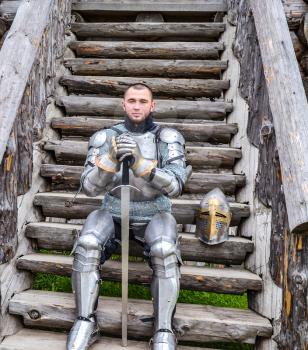 Knight in the armor on the wooden steps. Knightly armor and weapon. Semi - antique photo.