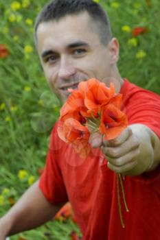 A young man in a red T-shirt is holding a bouquet of red poppies. The man is giving flowers. Poppy field.