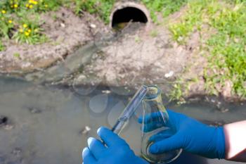 Check the water sample for infections. Hand in a glove with a bulb and a test tube against the background of a sewer pipe, dirty water. Pollution of ecology, environment