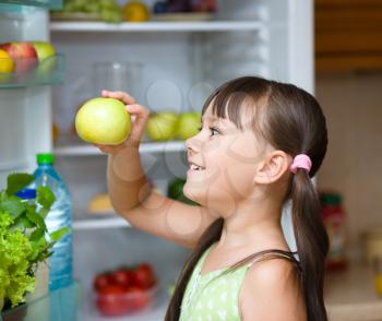 Happy girl eating apple standing near refrigerator with fruits and vegetables