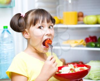 Happy girl eating salad standing near refrigerator with fruits and vegetables