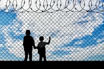 Refugee children concept. Silhouette of two children of refugees near the border fence of barbed wire