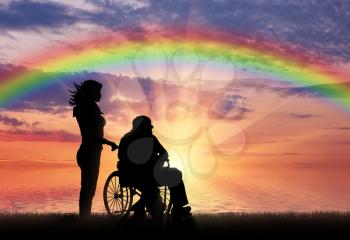 Silhouette of nurse caring for a disabled person in a wheelchair on a rainbow background. Concept of caring for a disabled person and house of the aged