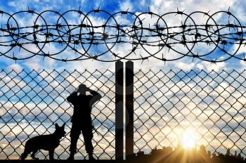 Safety concept. Silhouette of a military border guard with a dog on the background of the fence with barbed wire, and the city at sunset