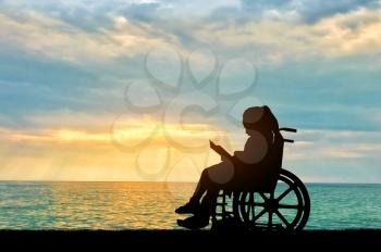 Silhouette of a disabled child girl sitting in a wheelchair reading a book on a sea sunset background. Conceptual image of the life of children with disabilities