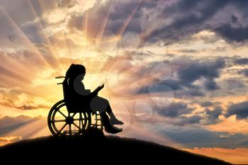 Silhouette of a disabled child girl sitting in a wheelchair reading a book on a sunset background on a hill. Life of children with disabilities