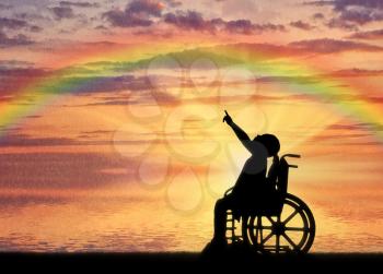Silhouette of a disabled child girl sitting in a wheelchair showing a finger at a rainbow against a sea sunset background. Conceptual image of the life of children with disabilities