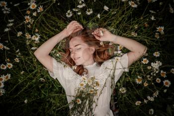 Beautiful woman enjoying daisy field, nice female lying down in meadow of flowers, pretty girl relaxing outdoor, having fun, holding plant, happy young lady and spring green nature, harmony concept