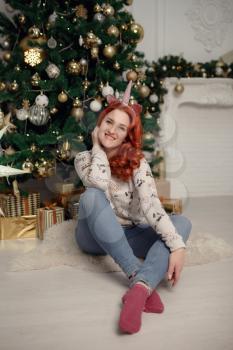 Attractive happy young woman in cozy comfortable clothes is sitting near a Christmas tree decorated with shiny toys among gold and red New Year's gifts. Magical festive atmosphere. Cute girl.
