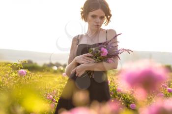 Beautiful young woman with curly hair posing near roses in a garden. The concept of perfume advertising.