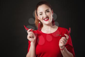 A chic red-haired young woman in a red overalls holds a hot chili pepper in her hands. Black background. Bright appearance.