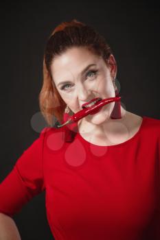 A chic red-haired young woman in a red overalls holds a hot chili pepper in her hands. Black background. Bright appearance.