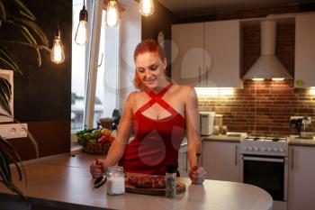 Photo of young pretty lady standing in kitchen while cooking meat. The sexy girl with a meat piece