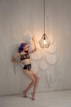 Beautiful young elegant dancer posing near an unusual lamp in her room. Delicate black lace underwear and purple hair