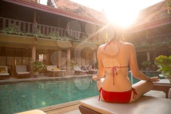 Young woman meditating near the pool at resort. soft beautiful light at sunset in a hotel in traditional Thai style, rest in an exotic. view from the back