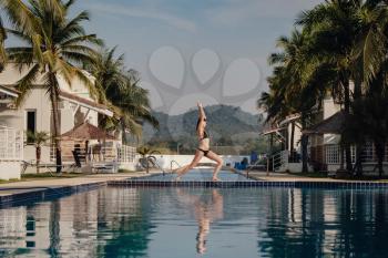 young woman practicing pose yoga on swimming pool and the nature beach at sunset or sunrise. Yoga and Healthy Concept. Warrior I or Virabhadrasana Pose